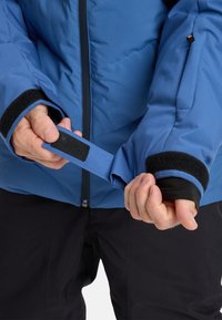 Person, der den Ärmel des blauen Jackets mit dem schwarzen Klettverschluss über der schwarzen Hose und dem Daumenlochärmel anpasst.