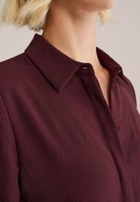 Camicia bordeaux con collo a punta, realizzata in tessuto leggero con una texture sottile e una caduta morbida.