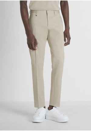 Uomo che indossa pantaloni beige a gamba dritta e sneaker bianche con i lacci, in piedi contro uno sfondo chiaro uniforme.