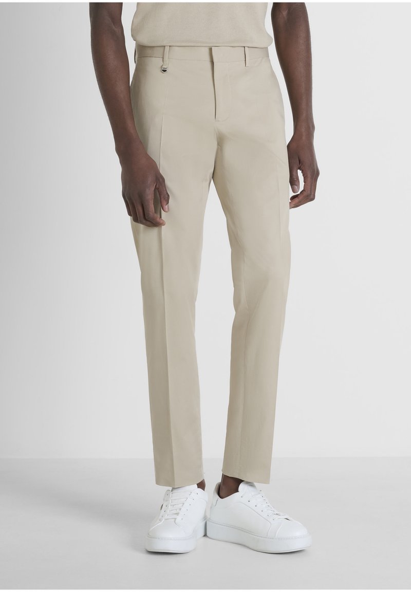 Homme portant un pantalon beige coupe droite et des baskets blanches à lacets, debout devant un fond clair uni.