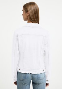 Chaqueta de mezclilla blanca con un dobladillo deshilachado, diseño estructurado y acentos de botones. Cuenta con mangas largas y un cuello clásico.