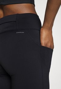 Črne legice z visokim pasom, z bočnim žepom. So izdelane iz elastičnega materiala z gladko teksturo in vidnimi šivi.