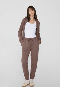 Brauner Kapuzenpullover und passende Hose aus weichem Stoff, mit einer Kordelzugtaille, Seitentaschen und elastischen Bündchen. Die Schuhe sind beige.