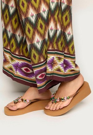 Piedi che indossano sandali con zeppa marroni adornati con pietre colorate, abbinati a tessuto fantasia con disegni geometrici verdi, marroni e viola.