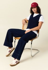 Marinblå stickad väst över en vit t-shirt, kombinerad med mörka slitna jeans och beige sneakers med vinröda accentuerade detaljer, sittande på en stol med flätad rygg.