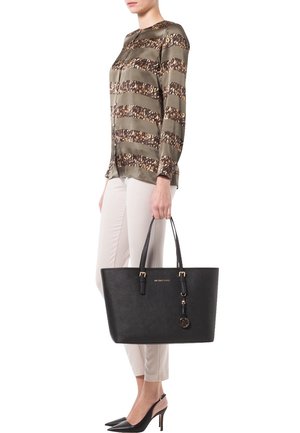 Mujer con pantalones beige y blusa marrón estampada sosteniendo un bolso grande negro con detalles dorados, que lleva zapatos de tacón negros puntiagudos.