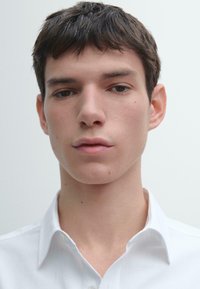 Primo piano di una persona con pelle chiara e capelli corti e scuri, che indossa una camicia bianca con colletto su uno sfondo neutro.