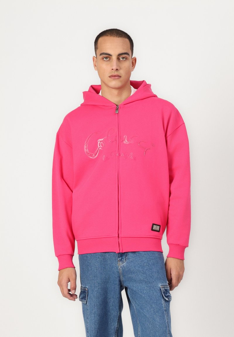 Sudadera de cremallera de color rosa brillante con capucha y un logo brillante en el frente. Presenta puños y dobladillo acanalados, combinada con pantalones de mezclilla.