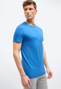 Άνδρας που φοράει φωτεινό μπλε κοντομάνικο μπλουζάκι και ανοιχτό γκρι παντελόνι, κοιτάζει προς τα αριστερά του πάνω σε ένα απλό λευκό φόντο.