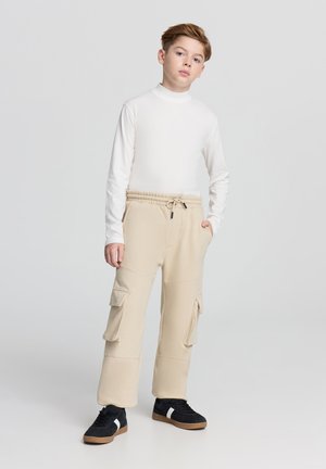 Pantaloni cargo beige con multiple tasche laterali, abbinati a un top bianco aderente a maniche lunghe. Scarpe da ginnastica nere con suola color cuoio e dettagli bianchi.