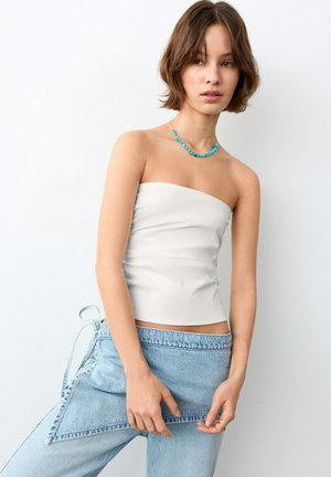 Junge Frau, die ein weißes trägerloses Oberteil und einen hellblauen Wickel-Jeansrock trägt, dazu eine Halskette aus türkisfarbenen Perlen als Accessoire.