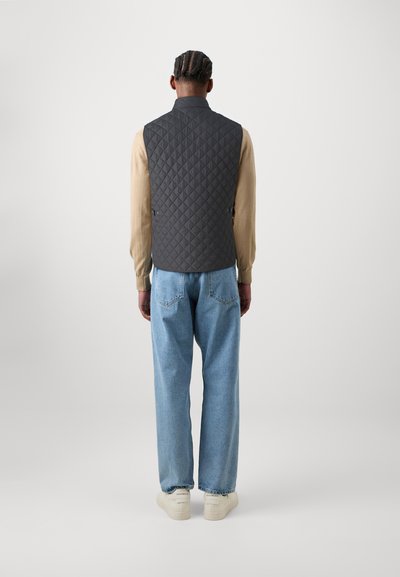 Gilet en tissu matelassé gris avec un col haut, associé à une chemise à manches longues beige et un jean bleu clair, porté avec des baskets blanches.