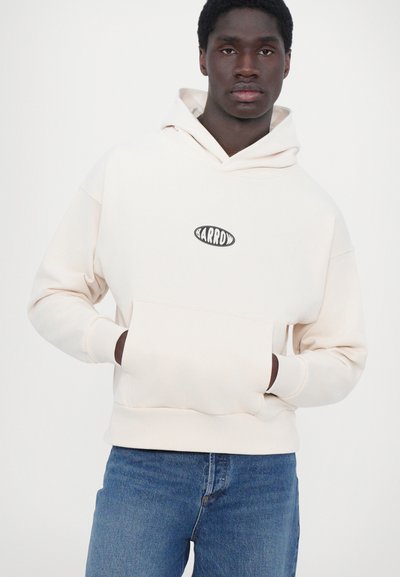 Ljusbeige hoodie i mjukt tyg, med en framficka i kängurustil och en svart oval logotyp på bröstet. Bärs med blå jeans.