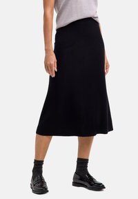 Donna che indossa una gonna nera midi, un top grigio, calze scure e mocassini neri lucidi, in piedi contro uno sfondo chiaro e uniforme.