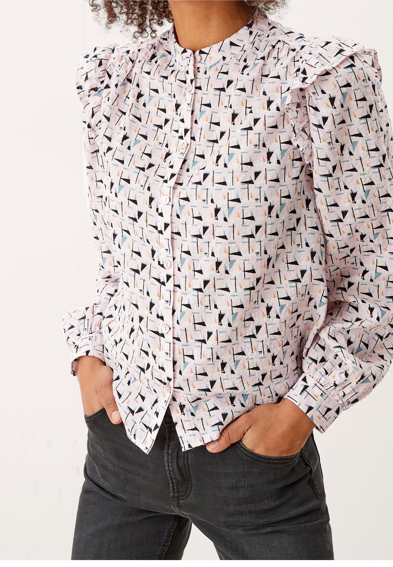 Femme portant une chemise blanche à boutons avec des motifs géométriques noirs, orange et turquoise, manches bouffantes, et un jean gris foncé, les mains dans les poches.