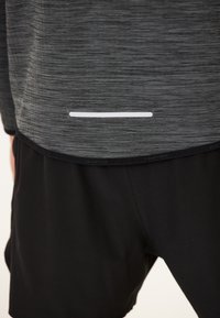 Grå, struktureret atletisk top med en vandret hvid stribe i kanten, parret med sorte shorts. Materialet virker let og åndbart.