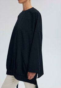 Osoba ubrana w luźny, ciemnoniebieski top z długim rękawem i marszczonym dołem, w połączeniu z jasnobeżowymi spodniami, stojąca na jednolitym tle.