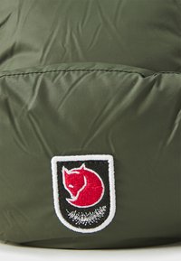 Olivengroen puffer stof met een gladde textuur, voorzien van een ronde patch met een rode vos-embleem op een zwart-witte achtergrond.