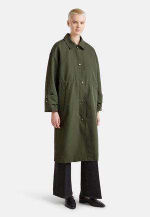 Imperméable vert olive avec une coupe décontractée, présentant un col, des boutons sur le devant et des poches latérales. Associé à un jean large noir.