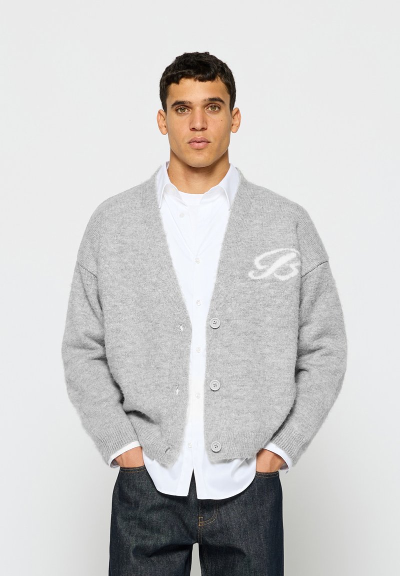 Šedý vlněný kardigan s uvolněným střihem, s V-výstřihem, zapínáním na knoflíky a bílým logem na hrudi. Nosený přes bílou košili.