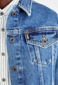 Veste en denim bleu clair avec un col classique, des accents en cuivre et des coutures orange. Elle présente des boutons-pression et un détail de poche poitrine.