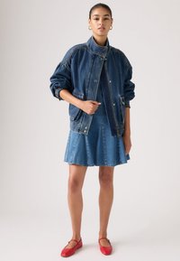 Νεαρή γυναίκα που φοράει υπερμεγέθη μπλε τζιν μπουφάν πάνω από τζιν φούστα και κόκκινα φλατ παπούτσια, στέκεται μπροστά σε απλό φόντο.