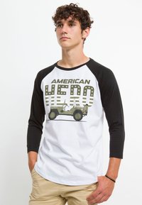 T-shirt raglan z czarnymi rękawami i białym korpusem, z napisem "AMERICAN HERO" oraz grafiką wojskowego jeepa w kolorze zielonym i szarym.