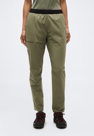 Person trägt olivgrüne Freizeithose und schwarze Wanderschuhe mit roten Schnürsenkeln, steht vor einem schlichten weißen Hintergrund.