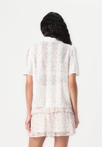 Top bianco all'uncinetto con maniche corte, caratterizzato da intricate trame in pizzo e una vestibilità ampia, abbinato a una gonna a strati con stampa floreale.