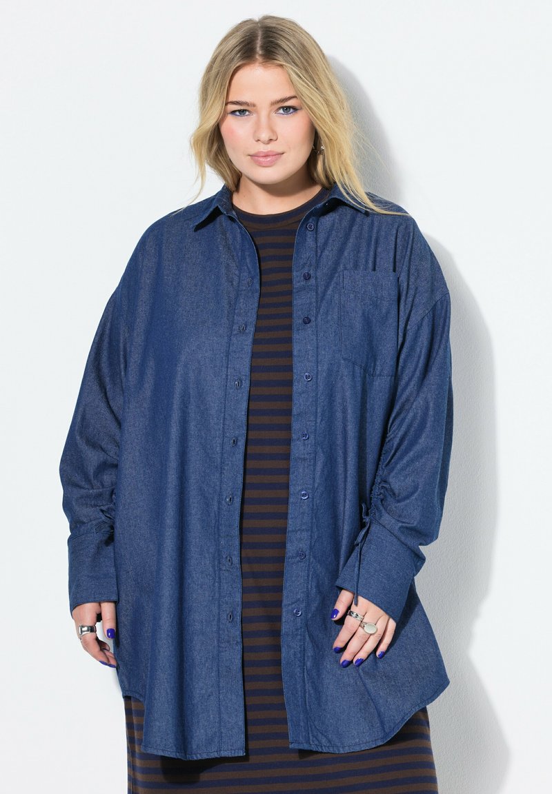 Camicia in denim blu con bottoni, maniche lunghe, vestibilità rilassata e tasca sul petto. Indossata sopra un vestito a righe, con dettagli sulle maniche arrotolate.