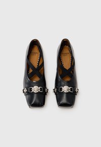 Black leather ballet flats with square toes, crossed elastic straps, and a silver decorative buckle on a strap with geometric accents.