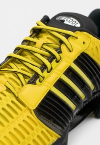 Κίτρινο αθλητικό παπούτσι με επάνω μέρος από δίχτυ, μαύρες λεπτομέρειες, ριγέ σχέδιο και κορδόνια, με υφή και εργονομικό σχεδιασμό.