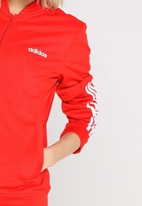 Veste à fermeture éclair rouge Adidas avec logo blanc sur la poitrine et trois bandes blanches sur les manches retroussées, main dans la poche latérale.