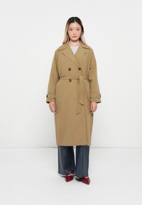 Cappotto trench beige con design doppiopetto, vita con cintura e bottoni grandi, abbinato a jeans a gamba larga e scarpe rosse.