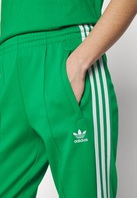 Green athletic pants made of smooth fabric featuring white side stripes and an "adidas" logo on the left thigh. Pockets on both sides.