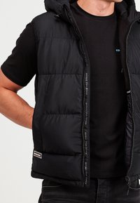 Gilet imbottito nero con cappuccio, dotato di chiusura a zip e pannelli cuciti. Logo accentuato sulla zip. Indossato sopra una maglietta nera.