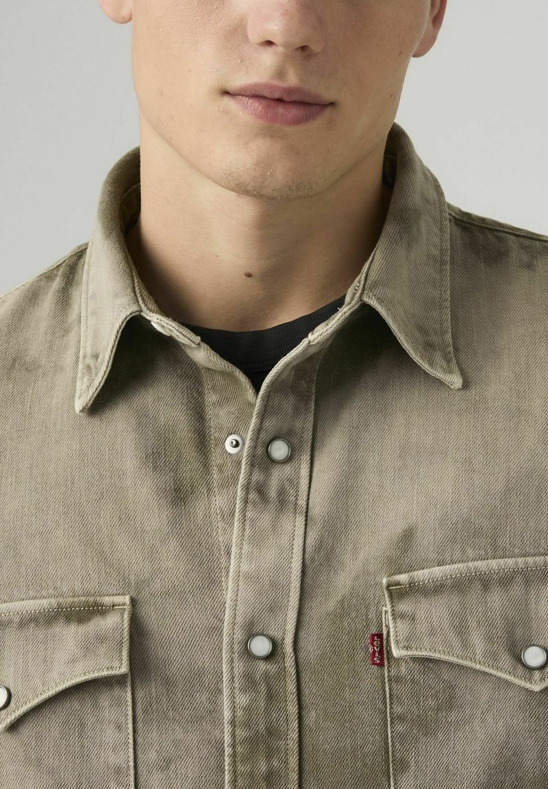Young man wearing a beige button-up shirt with two chest pockets and a small red Levi's tag, over a black undershirt.