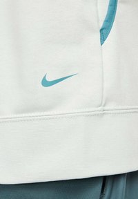 Hellgrauer Nike-Sweatshirt mit strukturiertem Finish, versehen mit einem kleinen blauen Logo. Das Design umfasst eine Fronttasche und kontrastierende blaue Akzente.