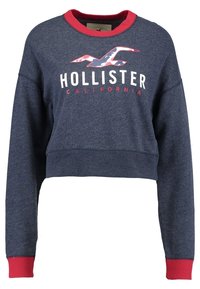 Zkrácená mikina v námořnické modři s červeným límcem a manžetami. Obsahuje grafické logo v bílé barvě a stylizovaný design ptáka. Měkká textura.