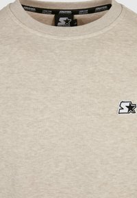 Sweat-shirt beige à col rond avec un petit logo étoile noir et blanc sur la poitrine et des étiquettes de la marque "Starter" à l'intérieur du col.