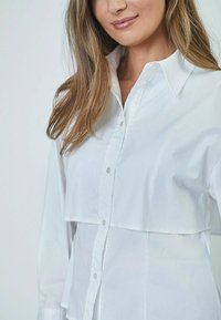 Camicia bianca con bottoni realizzata in tessuto morbido, caratterizzata da un design a strati con colletto e maniche lunghe, indossata da un modello con capelli lunghi.