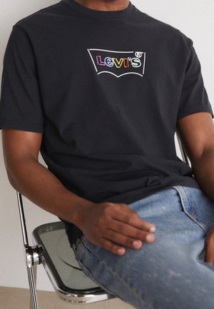 Man draagt een zwart Levi's T-shirt met een meerkleurig logo, zittend op een metalen stoel met een blauwe spijkerbroek.