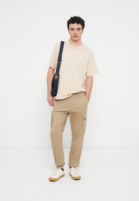 Μπεζ μπλουζάκι συνδυασμένο με χακί παντελόνι cargo με πλαϊνές τσέπες· ολοκληρωμένο με λευκά αθλητικά παπούτσια και μπλε σκούρη τσάντα ώμου.