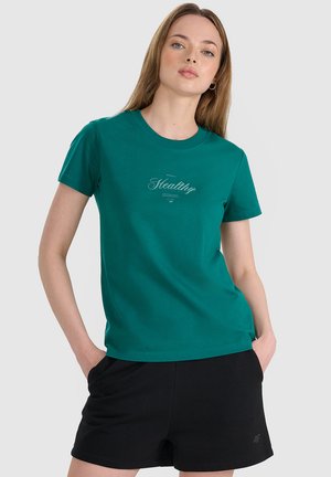 Kobieta o jasnej karnacji i długich włosach, ubrana w zielony t-shirt z napisem „Healthy” oraz czarne krótkie spodenki, z dłońmi w kieszeniach.