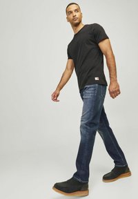 Tricou negru cu mânecă scurtă, asortat cu blugi drepti de culoare medie și pantofi negri cu talpă bej. Model plimbându-se pe un fundal simplu.