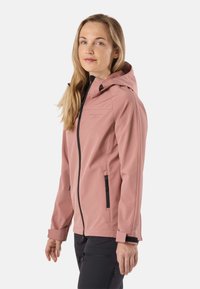 Moteris dėvi dulkinės rožinės spalvos gobtuvu puoštą minkštą striukę su juodu užtrauktuku ir juodas lauko kelnes, stovinčią prieš baltą tuščią foną.