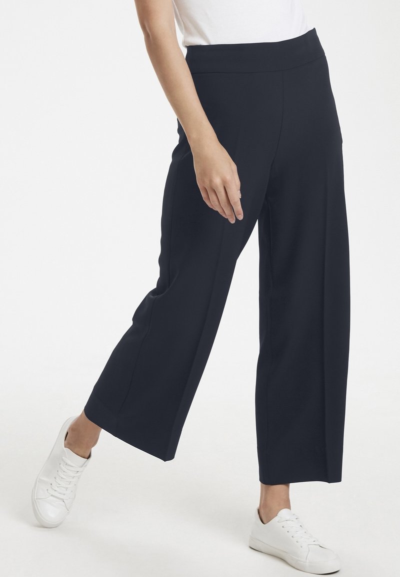 Navy wide-leg trousers in stretch fabric with a high waist, featuring flat front panels and side pockets, paired with white sneakers.