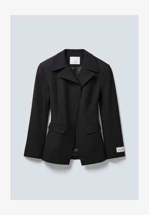 Melnā piegrieztā žakete ar platajiem lakatiem, divām priekšējām kabatām ar vāciņiem un zīmola etiķeti uz kreisā aproces, parādīta uz gaiši pelēka fona.