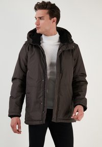 Brun isoleret jakke med fleece-forsynet hætte, knaplukning, sidelommer og en glat ydre tekstur. Båret over en grå sweater.