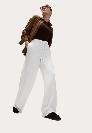 Donna in piedi con le mani nelle tasche, indossa una giacca marrone, una maglia nera, pantaloni bianchi a gamba larga, scarpe nere e occhiali da sole grandi.